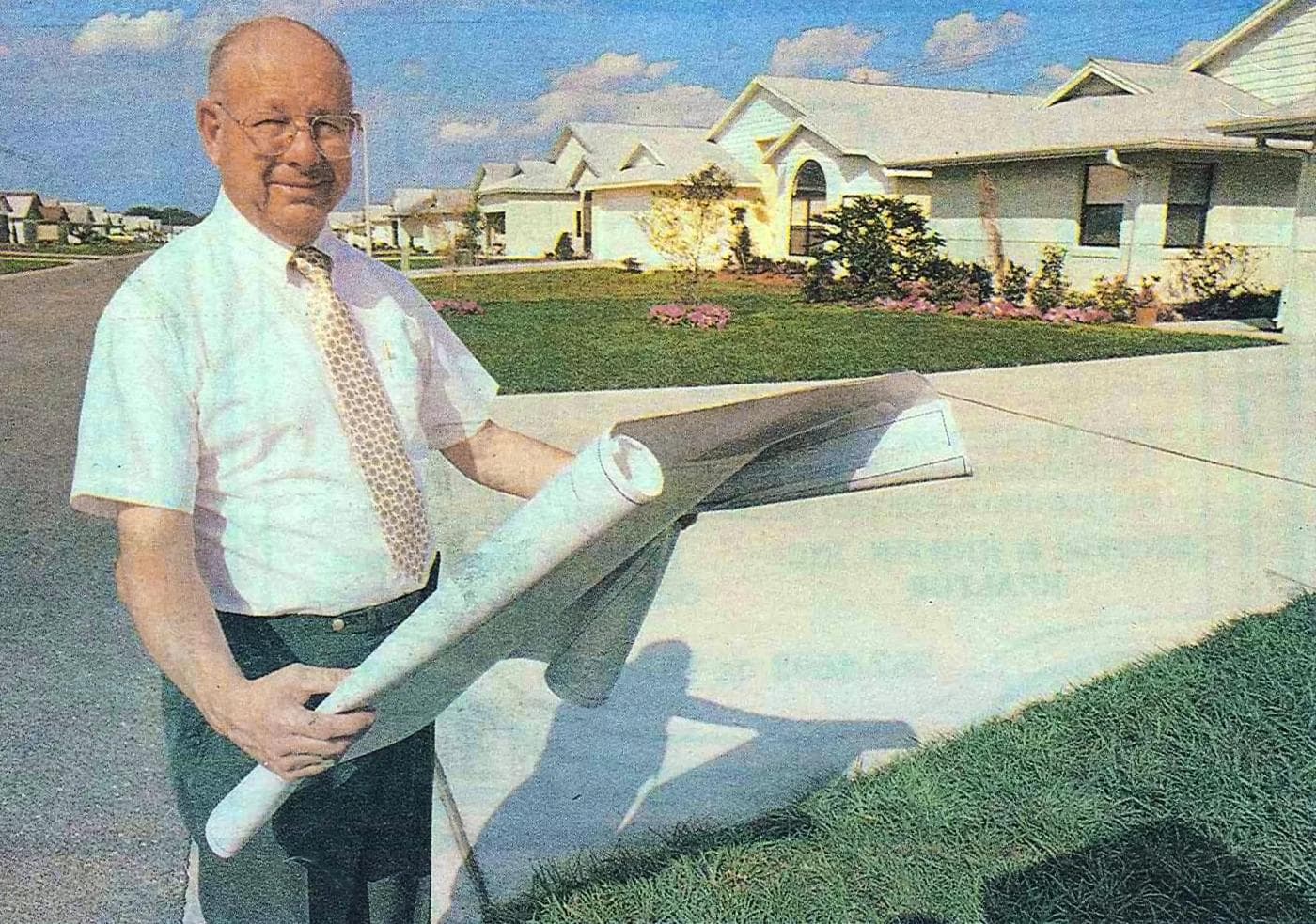 Sam Rodgers holding rolled blueprints in front of newly completed homes, 1990